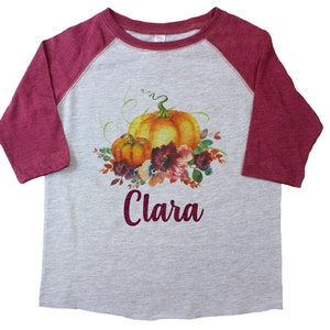 Pode incluir: Uma camiseta de beisebol cinza e bordô com um design de aquarela de abóboras e flores. O nome "Clara" está escrito em glitter bordô na frente da camiseta.