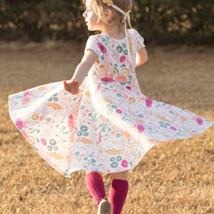 May include: A young girl wearing a white dress with a floral print and pink tights spins around in a field of dry grass.