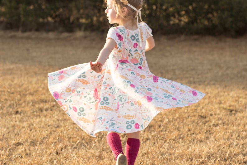 May include: A young girl wearing a white dress with a floral print and pink tights spins around in a field of dry grass.