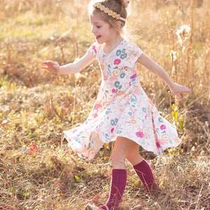 May include: A young girl wearing a white dress with a floral print and burgundy knee-high socks is twirling in a field of tall grass.