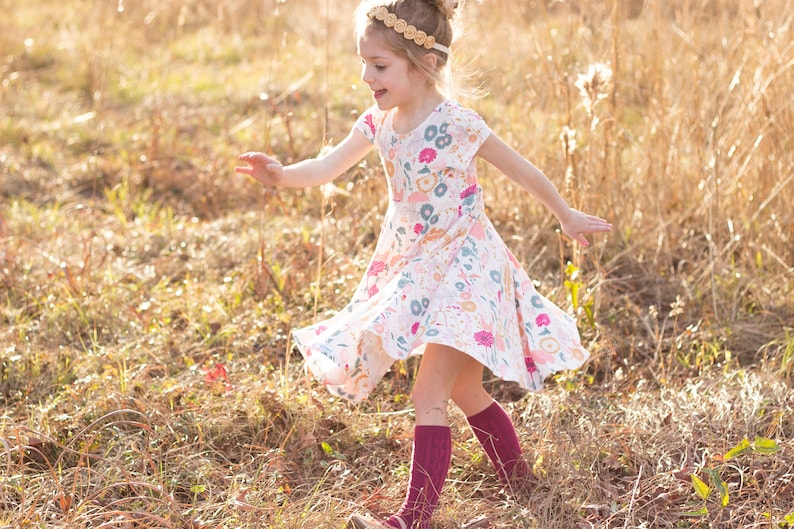 May include: A young girl wearing a white dress with a floral print and burgundy knee-high socks is twirling in a field of tall grass.