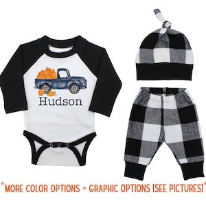 May include: A baby outfit set with a black and white buffalo plaid pattern. The set includes a long-sleeved baseball tee with a blue truck carrying pumpkins and the name "Hudson", black and white buffalo plaid pants, and a black and white buffalo plaid knotted hat.