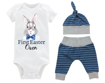 Puede incluir: Un body blanco para bebé con un conejito gris que lleva una pajarita azul. El body tiene el texto "First Easter Owen" impreso. El atuendo también incluye un gorro a rayas azul y gris y pantalones a juego.