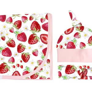 May include: A baby gift set with a pink and white swaddle blanket, a pink and white hat, and a pink and white headband. All items feature a watercolour strawberry print.