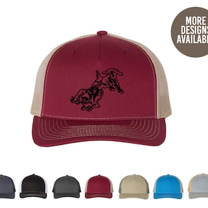 May include: A red and beige trucker hat with a black silhouette of two dogs playing. The hat has a mesh back and a curved brim.