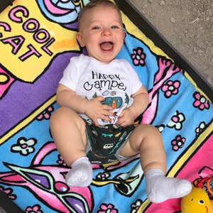 May include: A baby wearing a white onesie with the text "Happy Camper" and a cartoon camper van on it. The baby is lying on a colorful blanket with a cat design and the text "Cool Cat".