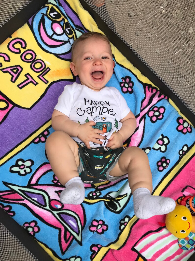May include: A baby wearing a white onesie with the text "Happy Camper" and a cartoon camper van on it. The baby is lying on a colorful blanket with a cat design and the text "Cool Cat".