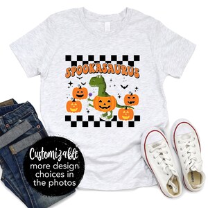 Może przedstawiać: Szary T-shirt z kratą i napisem "Spookasaurus" w pomarańczowych literach. Zielony dinozaur z dynią na głowie trzyma lizak. Wokół dinozaura znajdują się trzy dynie.