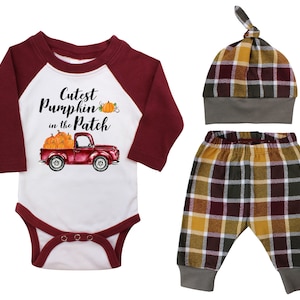 May include: A three-piece baby outfit set featuring a white long-sleeve bodysuit with a red truck carrying pumpkins and the text "Cutest Pumpkin in the Patch", a pair of plaid pants with brown, yellow, and white squares, and a matching plaid hat with a brown and yellow plaid pattern.