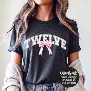 May include: A dark gray t-shirt with the word "TWELVE" in white letters with a pink bow. The shirt is being worn by a person with long brown hair.