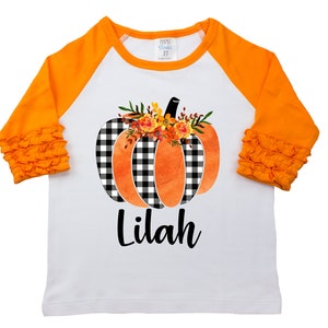 May include: White long-sleeved shirt with orange raglan sleeves and ruffles. The shirt features a black and white checked pumpkin with orange watercolour accents and a floral design. The name "Lilah" is printed below the pumpkin.