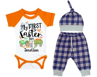 Puede incluir: Un conjunto para bebé niño para su primer Pascua, compuesto por un body blanco con mangas naranjas, un pantalón azul y blanco a cuadros y un gorro azul y blanco a cuadros. El body tiene el texto "My First Easter 2021 Jonathan" impreso.