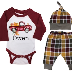 May include: A three-piece baby outfit set featuring a red truck with pumpkins, a burgundy and white long-sleeve shirt with the name "Owen", plaid pants in shades of brown, yellow, and burgundy, and a matching plaid hat with a grey brim.