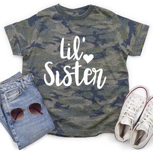 May include: A camouflage print t-shirt with the text "Lil' Sister" in white with a small white heart. The shirt is paired with blue jeans and white high-top sneakers.