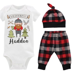 Op de afbeelding: Een driedelige baby-outfitset met een wit rompertje met een cartoon-houtzager-ontwerp, de tekst "WILDERNESS Haddon" en een rood-zwart ruitpatroon. De set bevat ook een bijpassende rood-zwart geruite muts en broek.