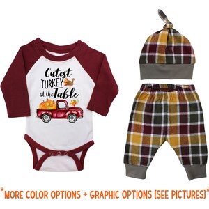 May include: A baby outfit set with a red and white long-sleeved onesie featuring a red truck with pumpkins and the text "Cutest Turkey at the Table". The set also includes a plaid hat and plaid pants.