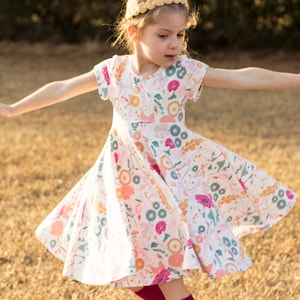 May include: A young girl wearing a white dress with a floral print and a gold headband. She is twirling in a field of dry grass.