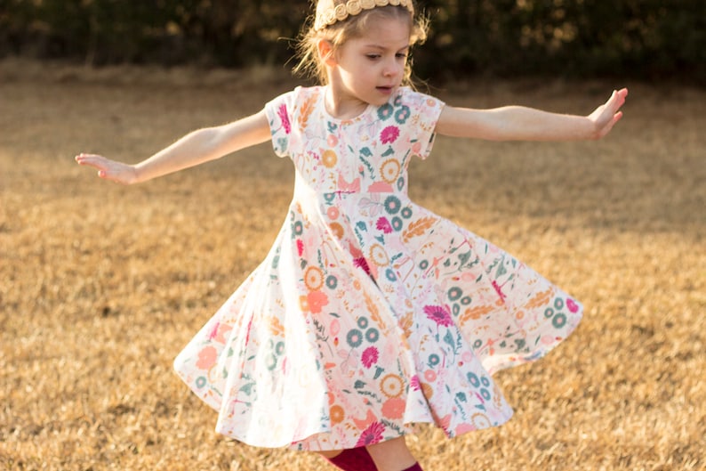 May include: A young girl wearing a white dress with a floral print and a gold headband. She is twirling in a field of dry grass.