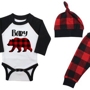 May include: A baby outfit set with a black and white long-sleeved onesie featuring a red and black plaid bear and the text "Baby", red and black plaid pants, and a red and black plaid knotted hat.