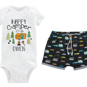 May include: A white baby onesie with the text "HAPPY CAMPER" and a cartoon camper van, trees, and a campfire. The onesie also has the name "OWEN" printed on it. The onesie is paired with black shorts with a pattern of colorful camper vans.
