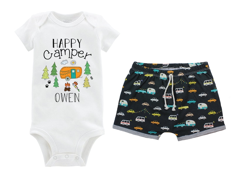 May include: A white baby onesie with the text "HAPPY CAMPER" and a cartoon camper van, trees, and a campfire. The onesie also has the name "OWEN" printed on it. The onesie is paired with black shorts with a pattern of colorful camper vans.