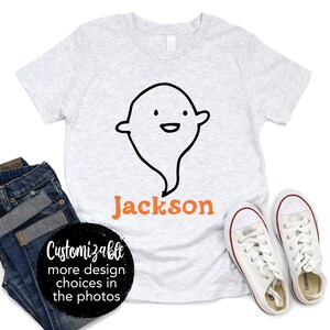 以下が含まれることがあります： 白いTシャツに、漫画の幽霊とオレンジ色の文字で「ジャクソン」と書かれています。このTシャツは、写真に掲載されているデザインからさらに多くのデザインを選んでカスタマイズできます。