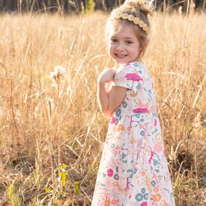 May include: A young girl wearing a white dress with a floral pattern and a brown headband. She is standing in a field of tall grass.