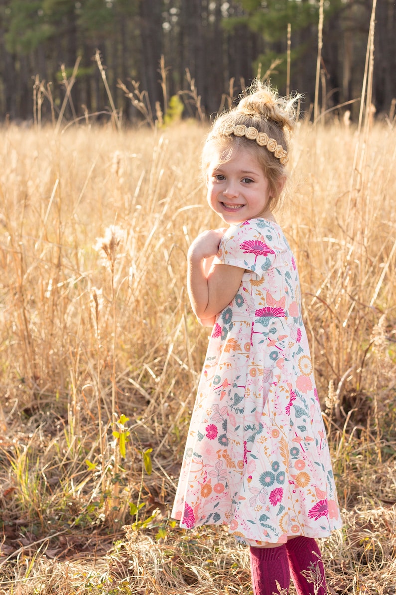 May include: A young girl wearing a white dress with a floral pattern and a brown headband. She is standing in a field of tall grass.