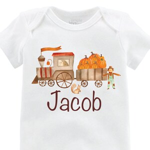 Könnte beinhalten: Ein weißer Baby-Strampler mit einem braunen Zug, der Kürbisse und einen Vogelscheuche transportiert. Der Zug hat ein rotes Dach und eine gelbe Flagge. Der Strampler ist mit dem Namen "Jacob" in braunen Buchstaben personalisiert.