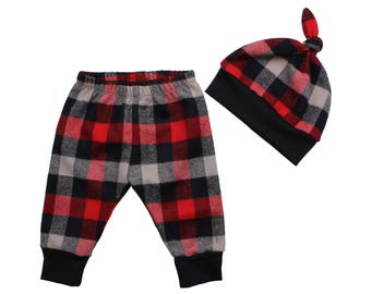 Op de afbeelding: Rode en zwarte geruite babybroek en bijpassende muts. De broek heeft een zwarte elastische tailleband en zwarte boorden. De muts heeft een zwarte rand en een geknoopte bovenkant.
