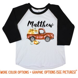 May include: A white and black baseball tee shirt with a red truck carrying pumpkins and the name "Matthew" printed on the front.