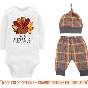 May include: A white long-sleeve bodysuit with a cartoon turkey graphic and the name "ALEXANDER" printed below. The outfit also includes a plaid hat and plaid pants.