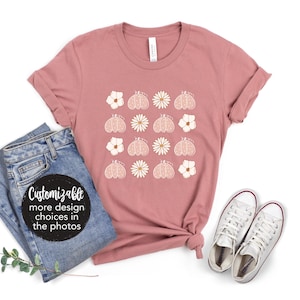 Pode incluir: Uma camiseta rosa pó com um padrão floral branco e borboleta. A camiseta é personalizável com mais opções de design disponíveis nas fotos.