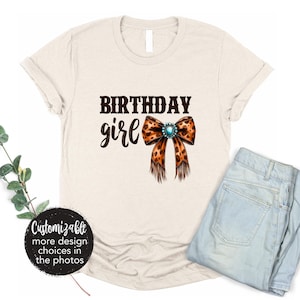 May include: A beige t-shirt with the text "Birthday girl" in brown lettering and a brown and orange leopard print bow with a turquoise jewel in the center.