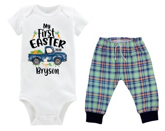 Puede incluir: Un body blanco para bebé con el texto "My First Easter 2020" y un camión azul con un conejito y zanahorias. El body se combina con pantalones de cuadros verdes y azules.