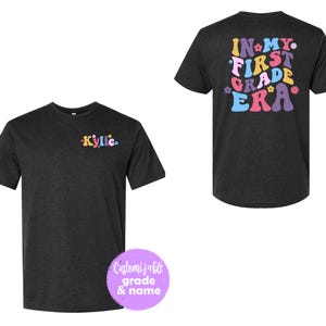 Puede incluir: Una camiseta negra con el texto "IN MY FIRST GRADE ERA" en letras de colores de estilo retro. El texto está rodeado de pequeños gráficos de flores. La camiseta es personalizable con un nombre y un grado.