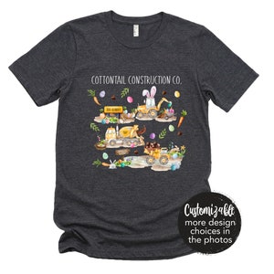 May include: A dark grey t-shirt with the text "Cottontail Construction Co." printed on the front. The design features a line of yellow construction vehicles with bunny ears and Easter eggs. The text "Big Bunny" is on a sign on one of the vehicles.