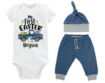 Puede incluir: Un body blanco para bebé con un camión azul que lleva zanahorias y un conejito. El body dice "My First Easter 2020" y "Bryson". El atuendo también incluye pantalones a rayas azules y blancas y un gorro a juego.
