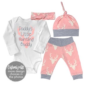 May include: A pink and white baby outfit with a white onesie that says "Daddy's Little Hunting Buddy" with a pink deer head graphic, a pink headband with a bow, pink and white patterned pants, and a pink and white hat with a grey brim.