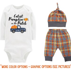 May include: A white baby bodysuit with long sleeves, featuring the text "Cutest Pumpkin in the Patch" and a blue truck carrying pumpkins. The set includes a plaid hat and pants with gray cuffs and waistband.