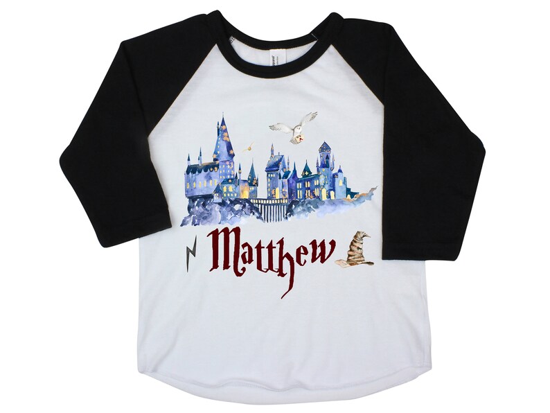 May include: A white and black baseball tee shirt with a watercolor illustration of Hogwarts castle, an owl flying overhead, and a Sorting Hat. The text "Matthew" is printed in red below the castle.
