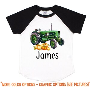 Puede incluir: Una camiseta de béisbol blanca y negra con un tractor verde y calabazas naranjas. La camiseta tiene el nombre "James" impreso en la parte delantera.