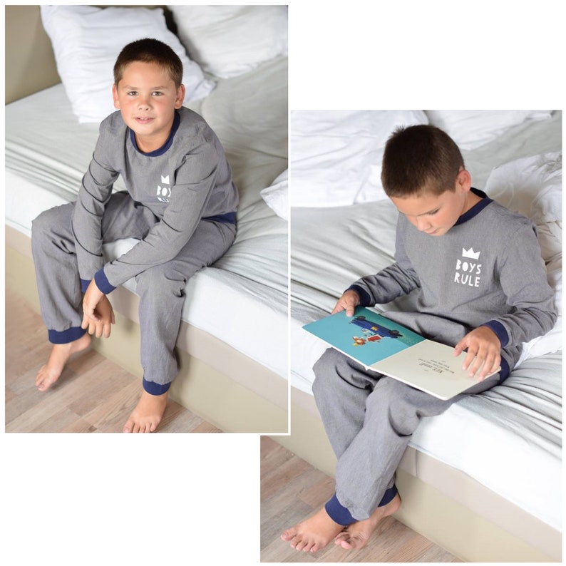 May include: A boy wearing gray pajamas with a blue trim and a white crown graphic that says "Boys Rule". He is sitting on a bed and reading a book.