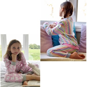 May include: Two young girls wearing pink and rainbow striped pajamas. One girl is sitting on a bed with a stuffed unicorn toy, while the other girl is sitting on a window seat looking out the window.