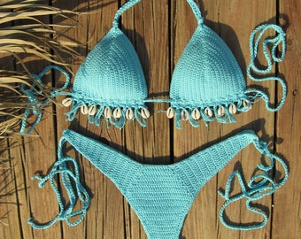 Puede incluir: Un conjunto de bikini de crochet azul claro con detalles de conchas blancas. La parte superior es de estilo triangular y la parte inferior es de estilo de corte alto.