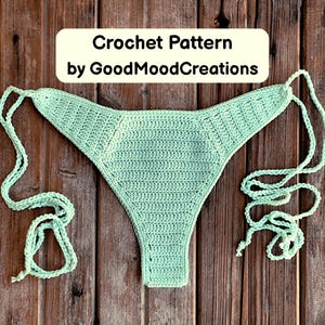 Puede incluir: Braguita de bikini de ganchillo verde menta con lazos, exhibida sobre una superficie de madera. El texto "Crochet Pattern by GoodMoodCreations" está en un rectángulo color crema encima de la braguita.