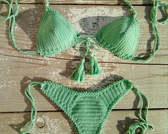 Puede incluir: Un top y una parte inferior de bikini de crochet verde con lazos de borla. El bikini está colocado sobre una superficie de madera clara.