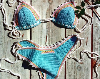 Puede incluir: Un conjunto de bikini de crochet en color azul claro con detalles en blanco, rosa y amarillo. La parte superior del bikini tiene forma de triángulo y la parte inferior es de estilo de cintura alta.
