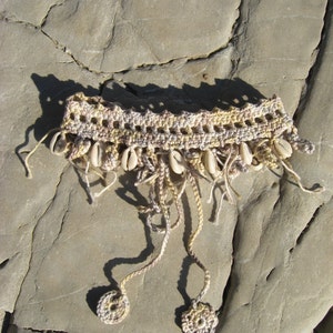 Puede incluir: Un collar tejido a crochet con hilo beige y marr&oacute;n y detalles de conchas blancas. El collar tiene dos borlas largas que cuelgan.