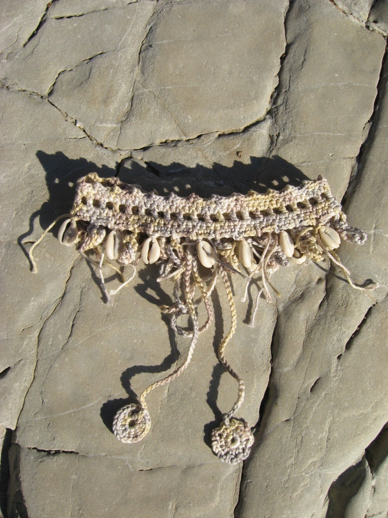 Puede incluir: Un collar tejido a crochet con hilo beige y marr&oacute;n y detalles de conchas blancas. El collar tiene dos borlas largas que cuelgan.